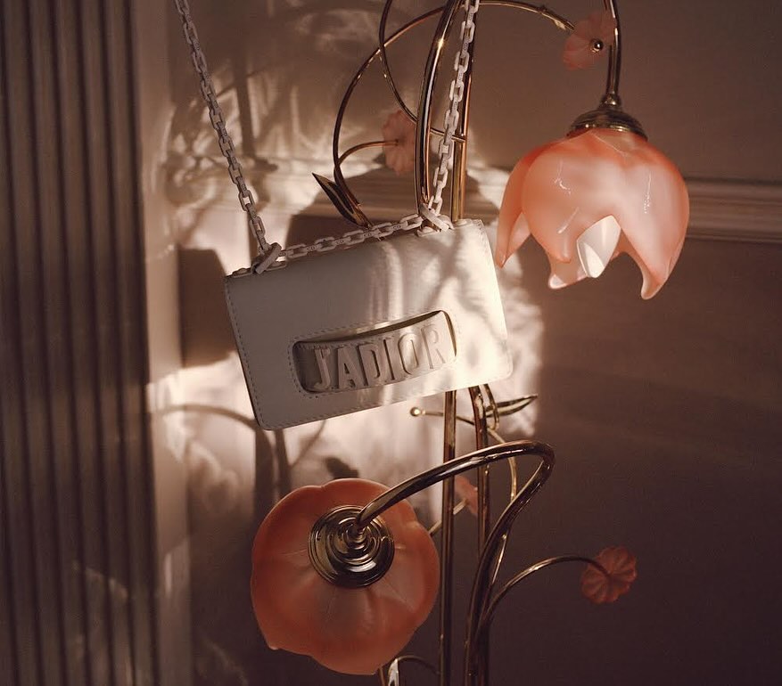 White leather handbag with a chain strap hanging from a gold-colored lamp with peach-colored glass shades, soft lighting, elegant.