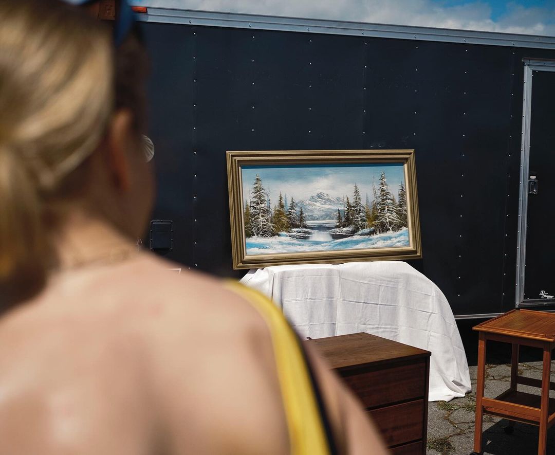 Painting of a snow-covered landscape on display outside near a black trailer, with a fair-skinned person in the foreground.