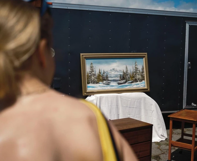 Painting of a snow-covered landscape on display outside near a black trailer, with a fair-skinned person in the foreground.