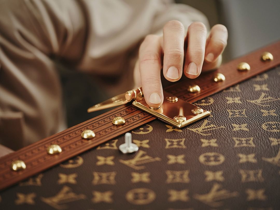 Close-up of person's hand adjusting hardware on a brown designer trunk with gold accents, showcasing craftsmanship and detail.