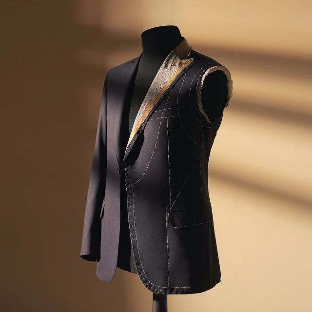Unfinished dark blue suit jacket on a black mannequin stand against a beige wall with window shadow patterns for tailoring work.