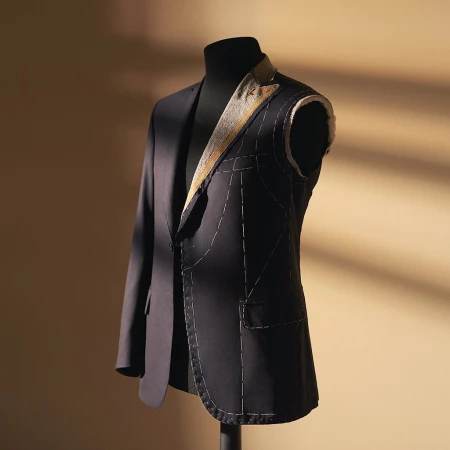 Unfinished dark blue suit jacket on a black mannequin stand against a beige wall with window shadow patterns for tailoring work.