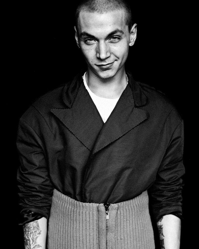 Monochrome portrait of a young adult male with short hair, wearing a dark jacket, against a stark black background. Close-up studio shot.