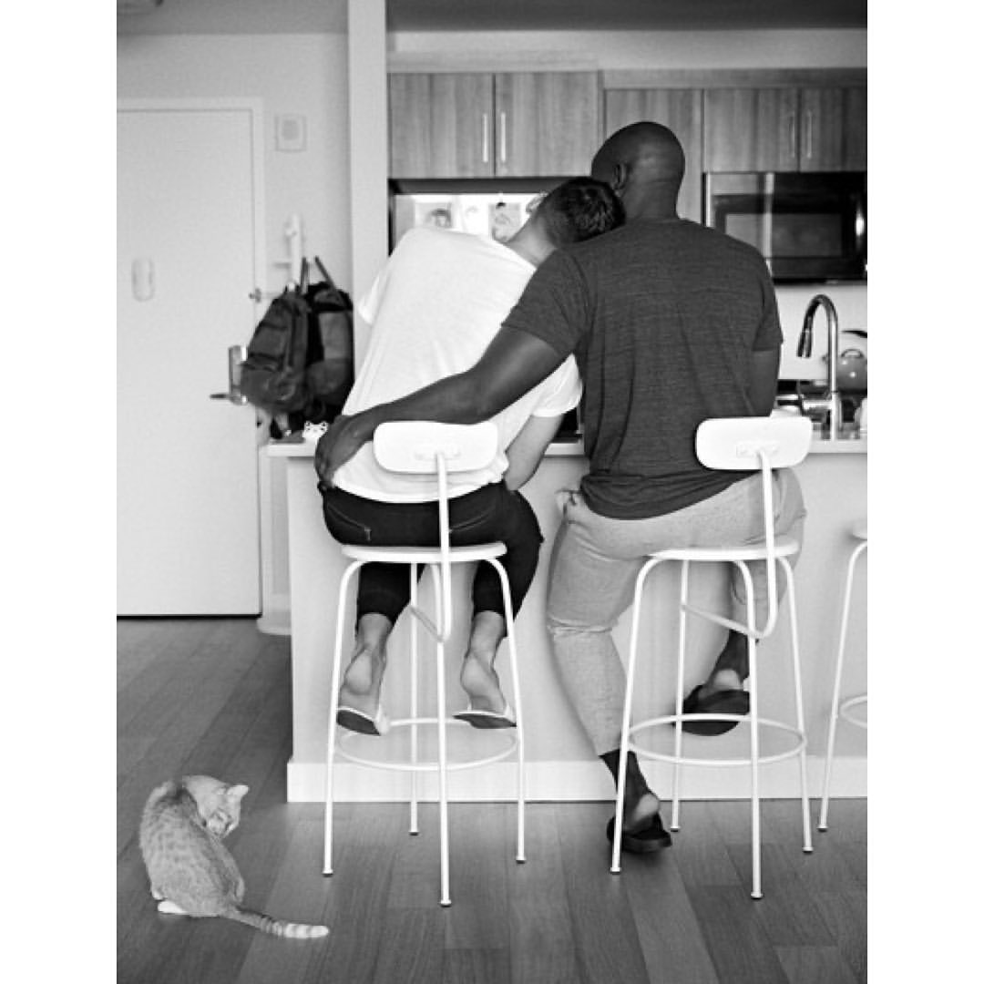 Two adults sit on stools at a counter in an indoor kitchen with a cat on the floor, showcasing a domestic scene in black and white.
