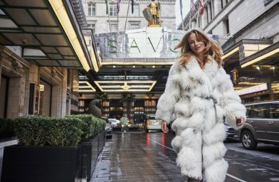 Woman in a long white fluffy fur coat walking outside The Savoy hotel entrance in London, photographed by Chris Craymer.