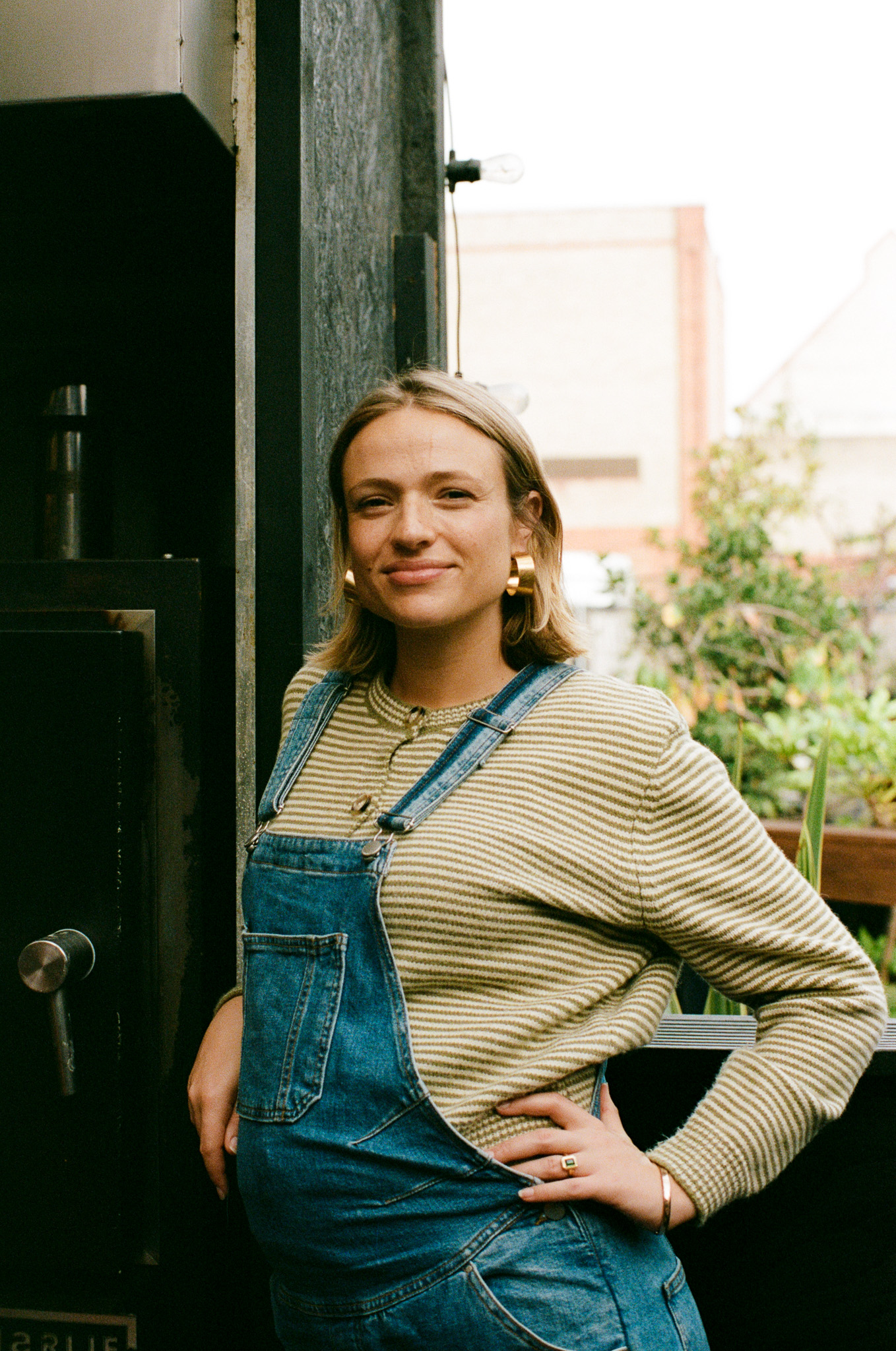 A woman poses in denim overalls and a striped shirt in front of a dark grey wall with green foliage. She is smiling and wearing gold earrings.