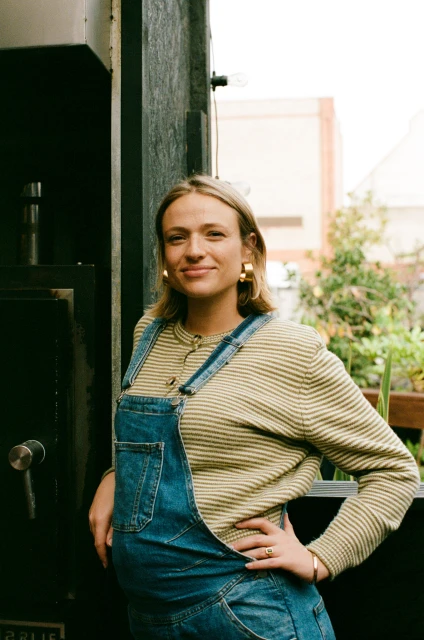A woman poses in denim overalls and a striped shirt in front of a dark grey wall with green foliage. She is smiling and wearing gold earrings.