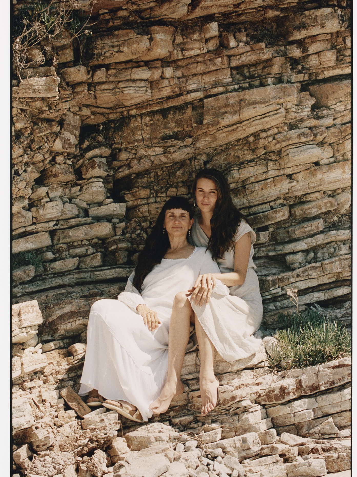 Two women pose outdoors against a rocky backdrop. The woman in the foreground is seated, wearing a long white dress. The background is blurred.