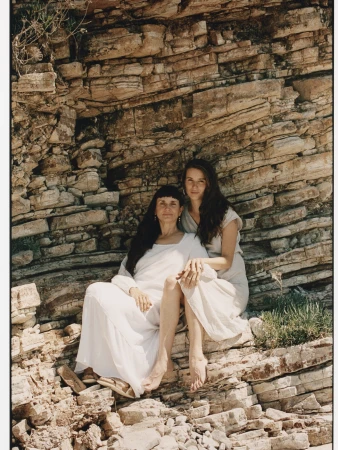 Two women pose outdoors against a rocky backdrop. The woman in the foreground is seated, wearing a long white dress. The background is blurred.