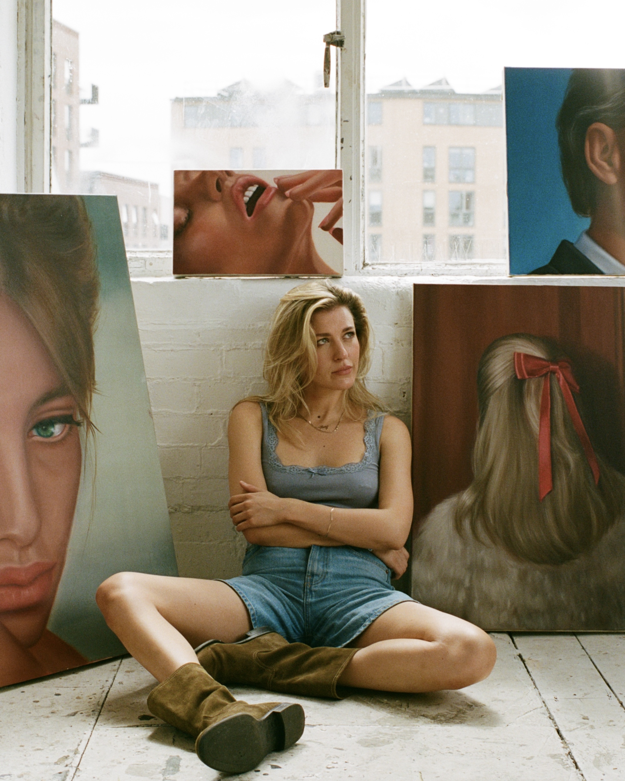 Blonde woman in denim shorts and brown boots sits surrounded by paintings in a bright, airy studio with artwork leaning against the wall.