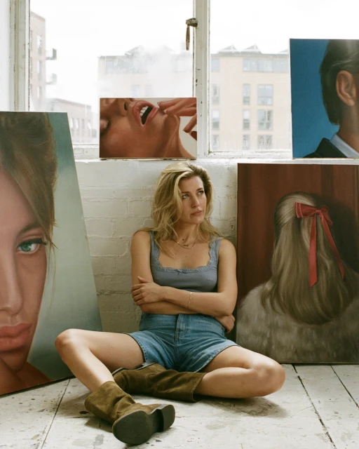 Blonde woman in denim shorts and brown boots sits surrounded by paintings in a bright, airy studio with artwork leaning against the wall.