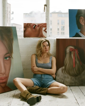 Blonde woman in denim shorts and brown boots sits surrounded by paintings in a bright, airy studio with artwork leaning against the wall.
