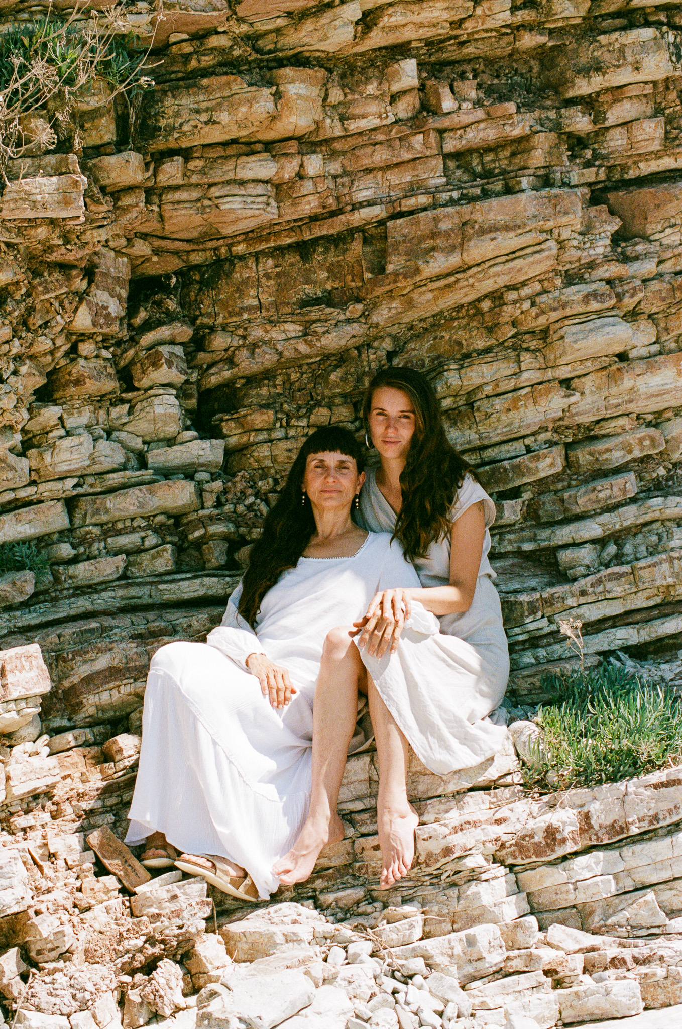 Two women in flowing dresses pose on a rocky outcrop with layered rock formations in the background on a sunny day.