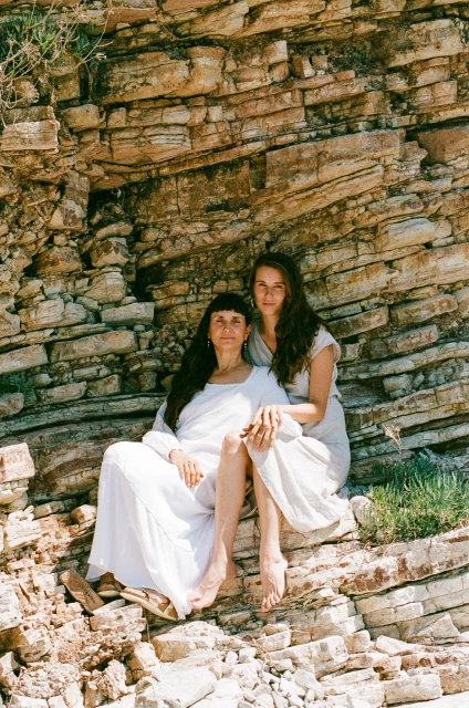 Two women in flowing dresses pose on a rocky outcrop with layered rock formations in the background on a sunny day.