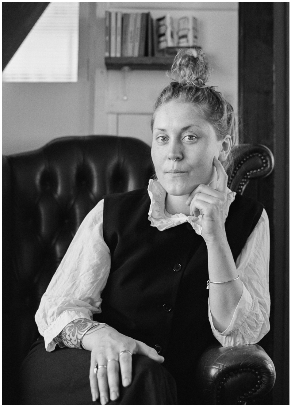 Black and white portrait of a woman with an updo hairstyle sitting in a leather chair in a well-lit indoor setting.