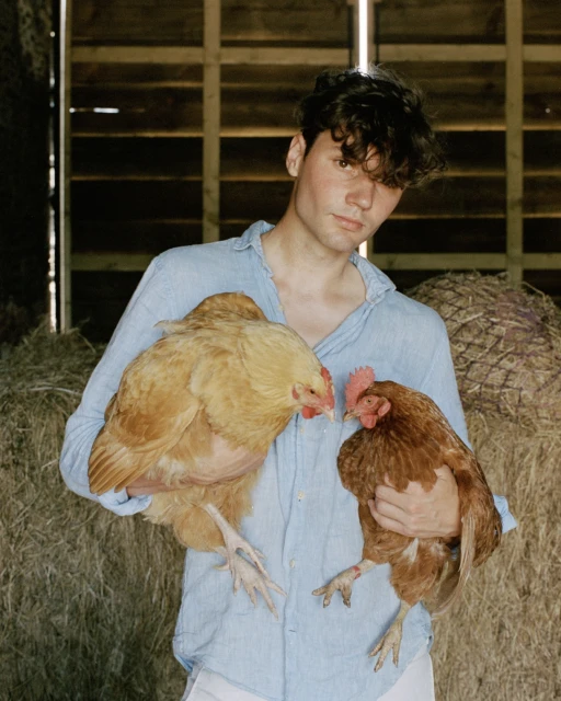 Young man in a blue shirt holds two hens against a background of hay bales, creating a rural and intimate portrait.