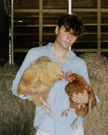 Young man in a blue shirt holds two hens against a background of hay bales, creating a rural and intimate portrait.