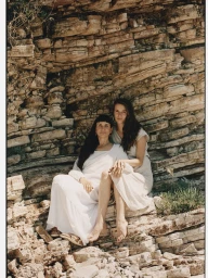 Two women in white dresses seated together on a rocky cliffside ledge outdoors.