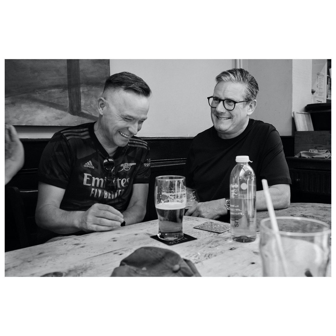 Two men in pub setting wearing jerseys, sitting at table with beer and water bottle, sharing a light moment, captured in black and white.