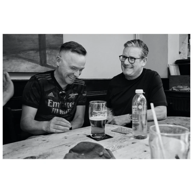 Two men in pub setting wearing jerseys, sitting at table with beer and water bottle, sharing a light moment, captured in black and white.