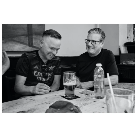 Two men in pub setting wearing jerseys, sitting at table with beer and water bottle, sharing a light moment, captured in black and white.