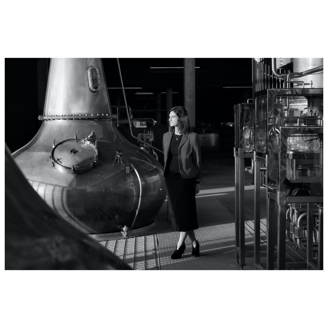 A black and white shot of a woman standing next to a large copper distillery still in a modern industrial setting.