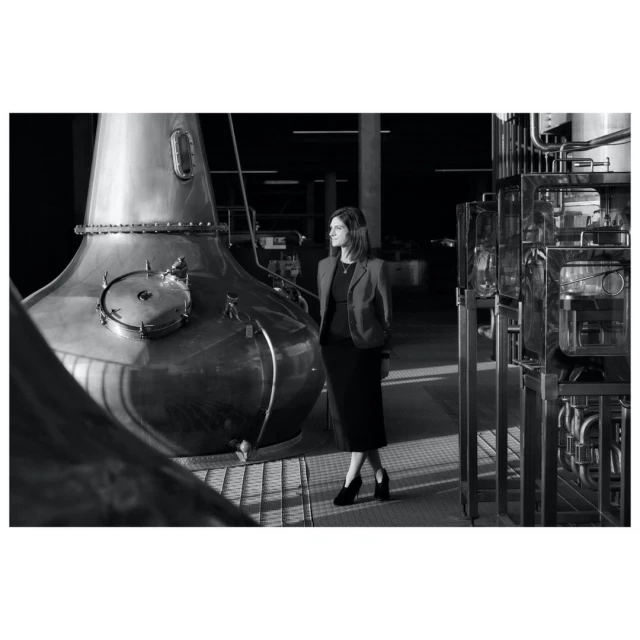 A black and white shot of a woman standing next to a large copper distillery still in a modern industrial setting.