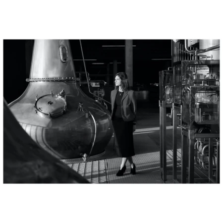 A black and white shot of a woman standing next to a large copper distillery still in a modern industrial setting.