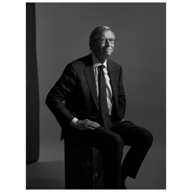 Black and white portrait of an adult male wearing a suit, tie, and glasses, sitting on a box in an indoor setting with a grey background.