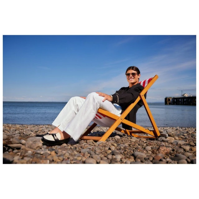 Adult in sunglasses relaxes in a beach chair on pebble beach against a backdrop of blue sky and ocean.