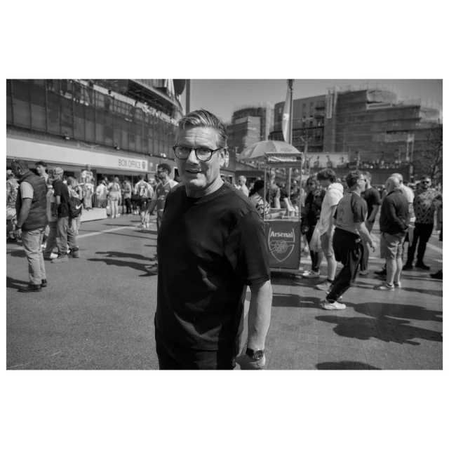 Black and white shot of a man in glasses and a dark shirt standing in front of a crowd of people outside of a building.