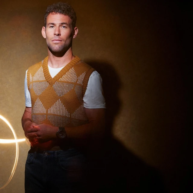 Portrait of a man with curly brown hair wearing a yellow patterned sweater vest, jeans, and a watch in a dimly lit indoor studio.
