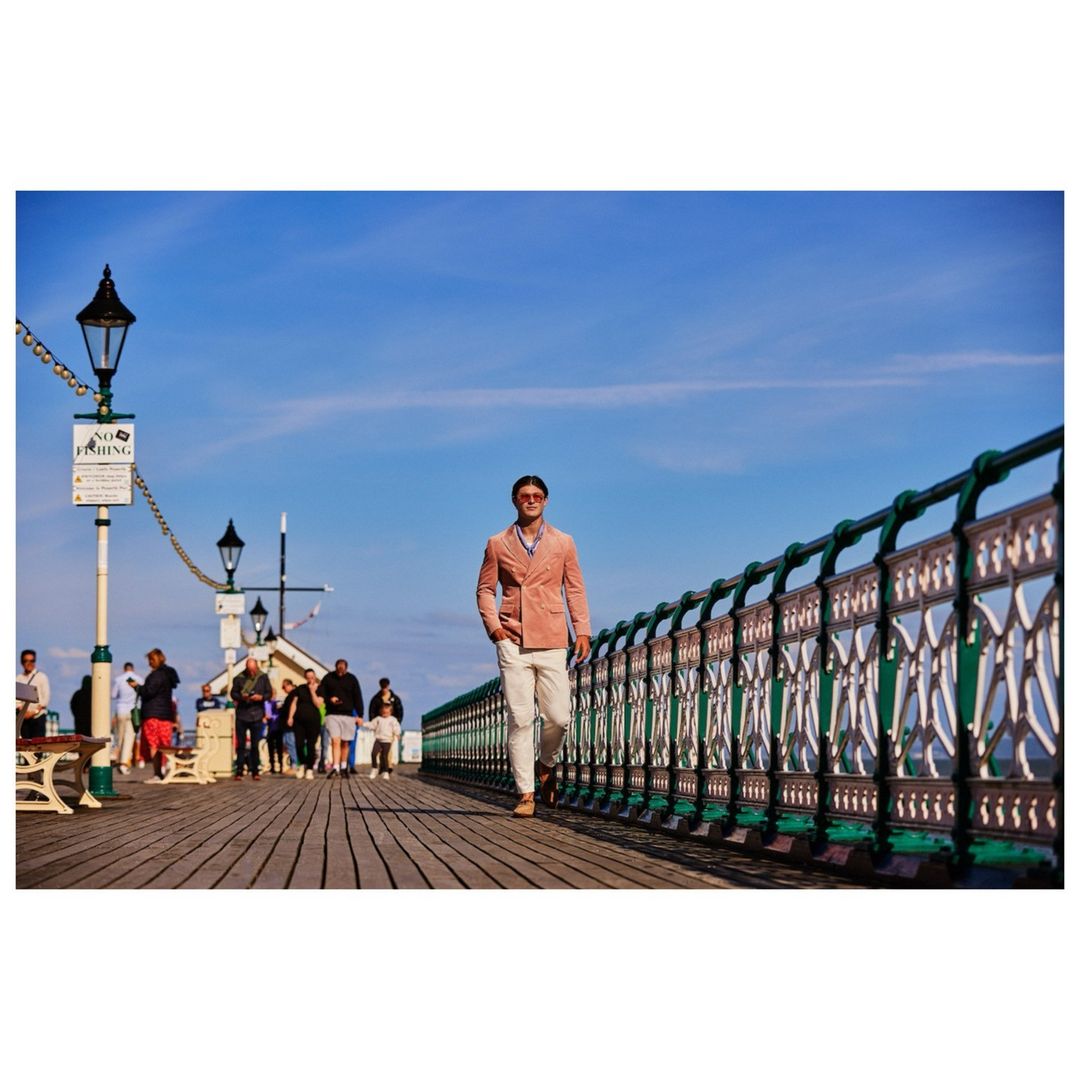 Man in a coral jacket walking along a seaside pier on a sunny day, with a view of the sky, the sea, and other people in the background.