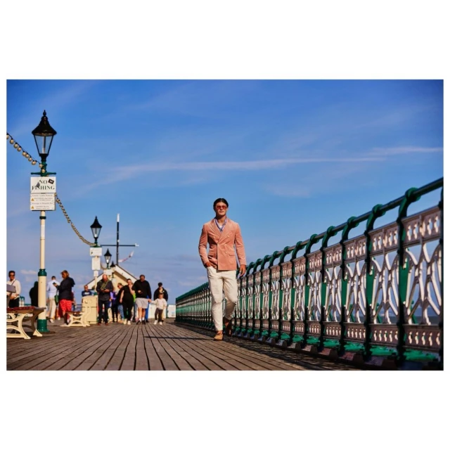 Man in a coral jacket walking along a seaside pier on a sunny day, with a view of the sky, the sea, and other people in the background.