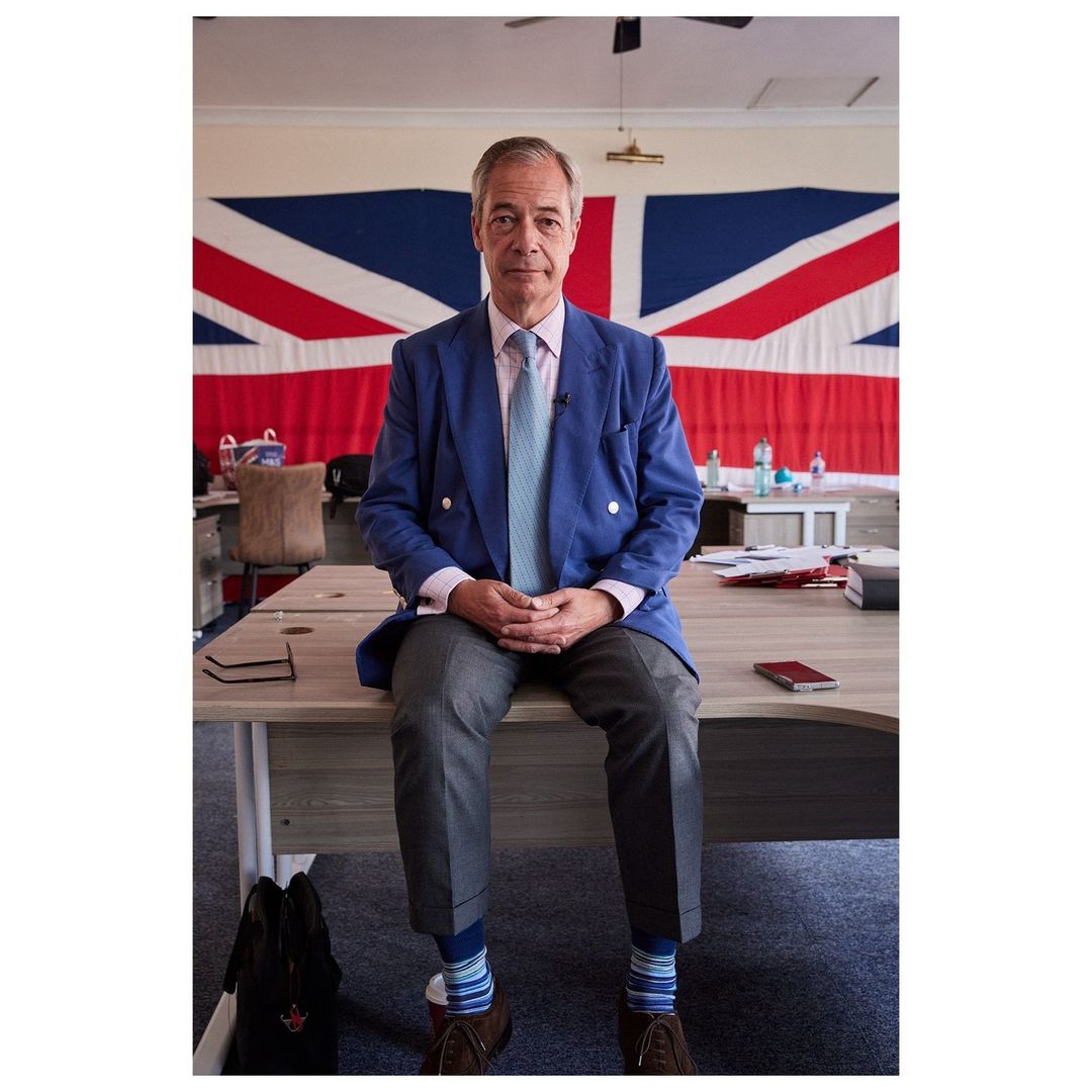 Adult male in blue jacket sits on desk with United Kingdom flag in background in indoor setting with natural lighting.