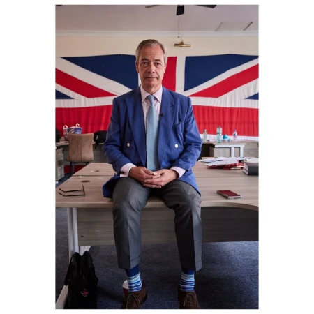 Adult male in blue jacket sits on desk with United Kingdom flag in background in indoor setting with natural lighting.