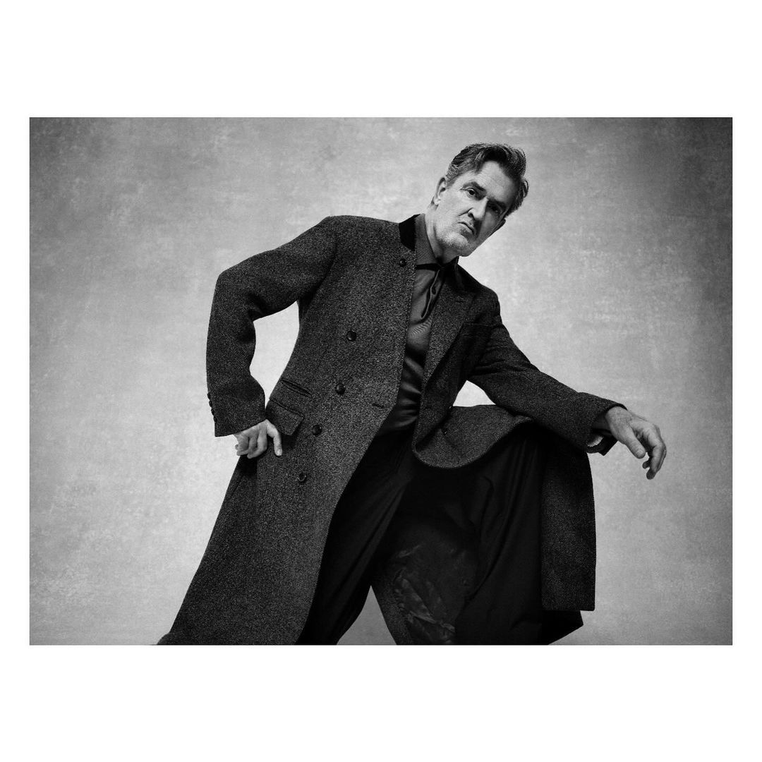 Black-and-white portrait of a mature adult man in a coat and wide-legged pants, posing in a studio setting, looking at the camera.