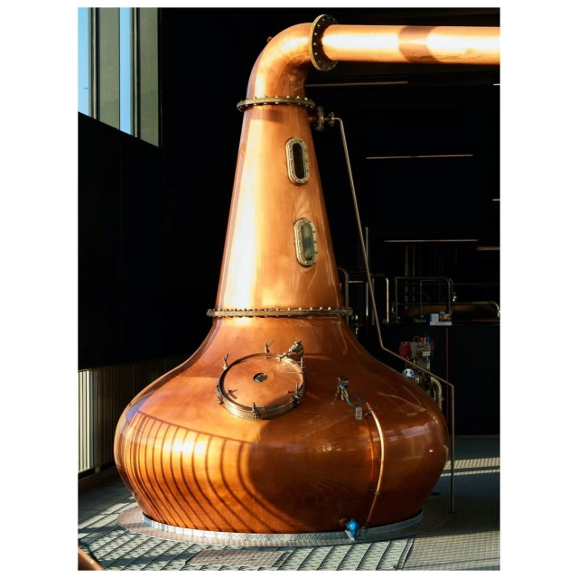 Large copper pot still inside a distillery, used for spirits production, lit by natural sunlight streaming from nearby windows.