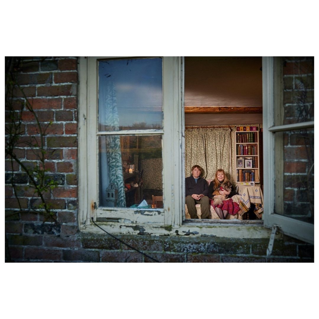 Man, girl, and dog posed in a rural window with a brick exterior, a landscape view, and indoor furnishings in natural light.