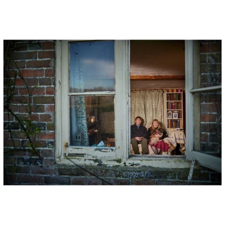 Man, girl, and dog posed in a rural window with a brick exterior, a landscape view, and indoor furnishings in natural light.