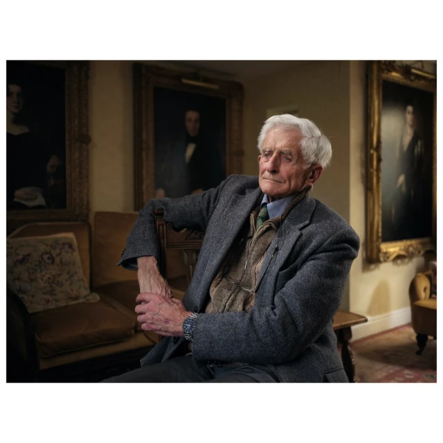 Elderly man with white hair sits indoors with arms crossed, wearing a tweed jacket; portraits are hung in the background.