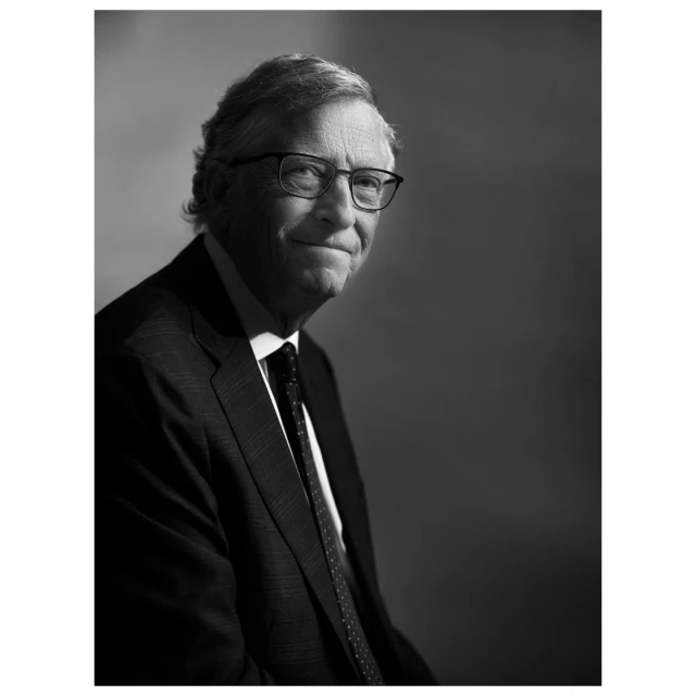 Black and white portrait of an adult male with glasses wearing a suit and tie against a blurred grey background in soft studio lighting.