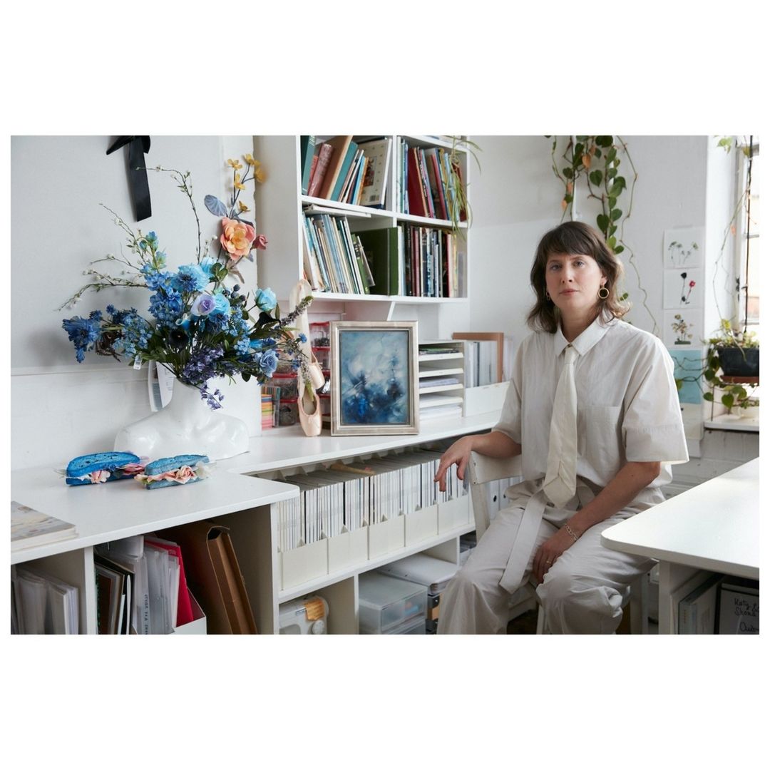 Woman in white shirt and pants sits by white desk adorned with flowers, books, art, and ballet slippers in a bright, natural light.