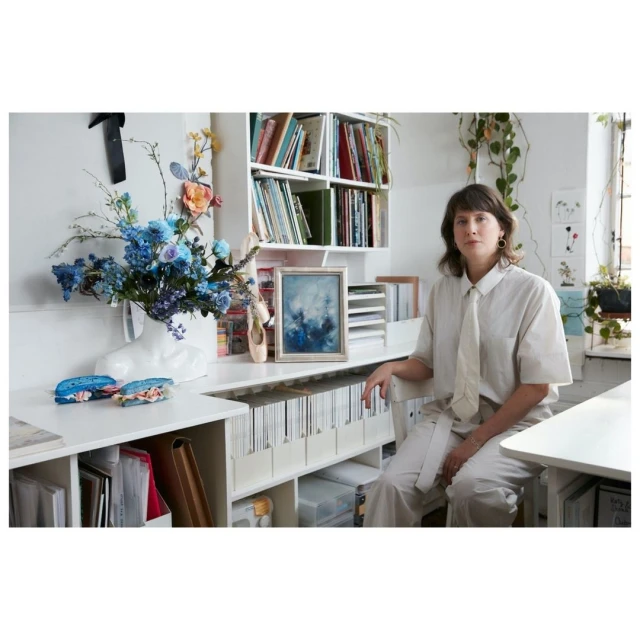 Woman in white shirt and pants sits by white desk adorned with flowers, books, art, and ballet slippers in a bright, natural light.