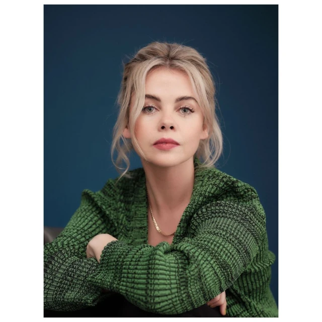 Portrait of a fair-skinned blonde woman with her arms crossed, wearing a green cardigan against a dark blue background. 