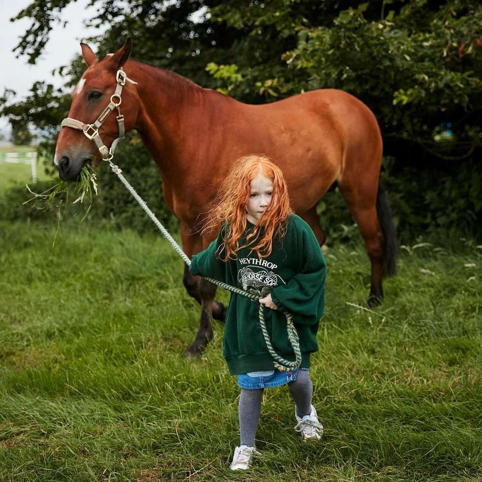 A young red-haired girl leads a large brown horse in a grassy field with trees in the background on a cloudy day.