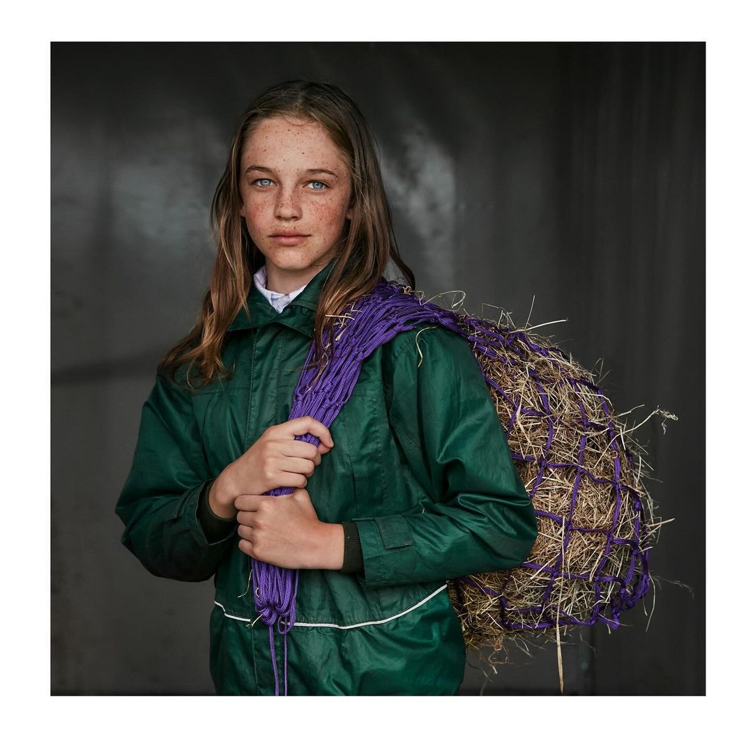 Portrait of a freckled preteen girl in a green jacket, carrying a bale of hay wrapped in purple netting over her shoulder.