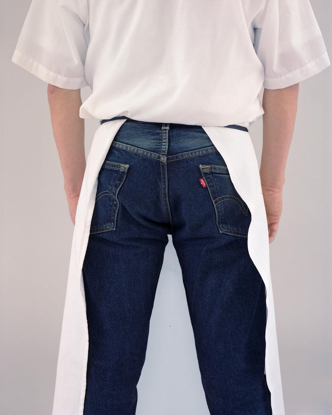 Rear view of a person in blue jeans wearing a white apron and white short-sleeved shirt, against a plain white background.