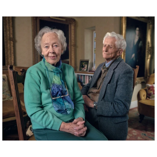 Portrait of elderly woman and man in a vintage-styled living room. Woman seated in front, man seated behind her. Warm indoor lighting.