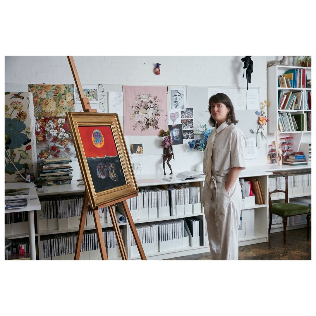 An artist stands next to a wood easel displaying her painting with a red and black color palette inside an art studio.